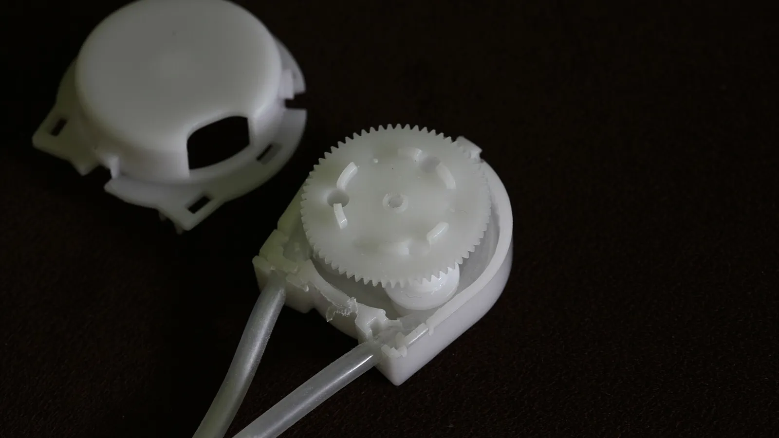 White plastic peristaltic pump head with the cover removed, exposing the toothed rotor, roller and curved tube track on a dark background